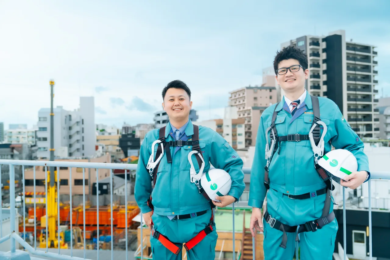建設現場を背景にヘルメットを手に笑顔で並ぶ安全帯を着けた若手社員2名